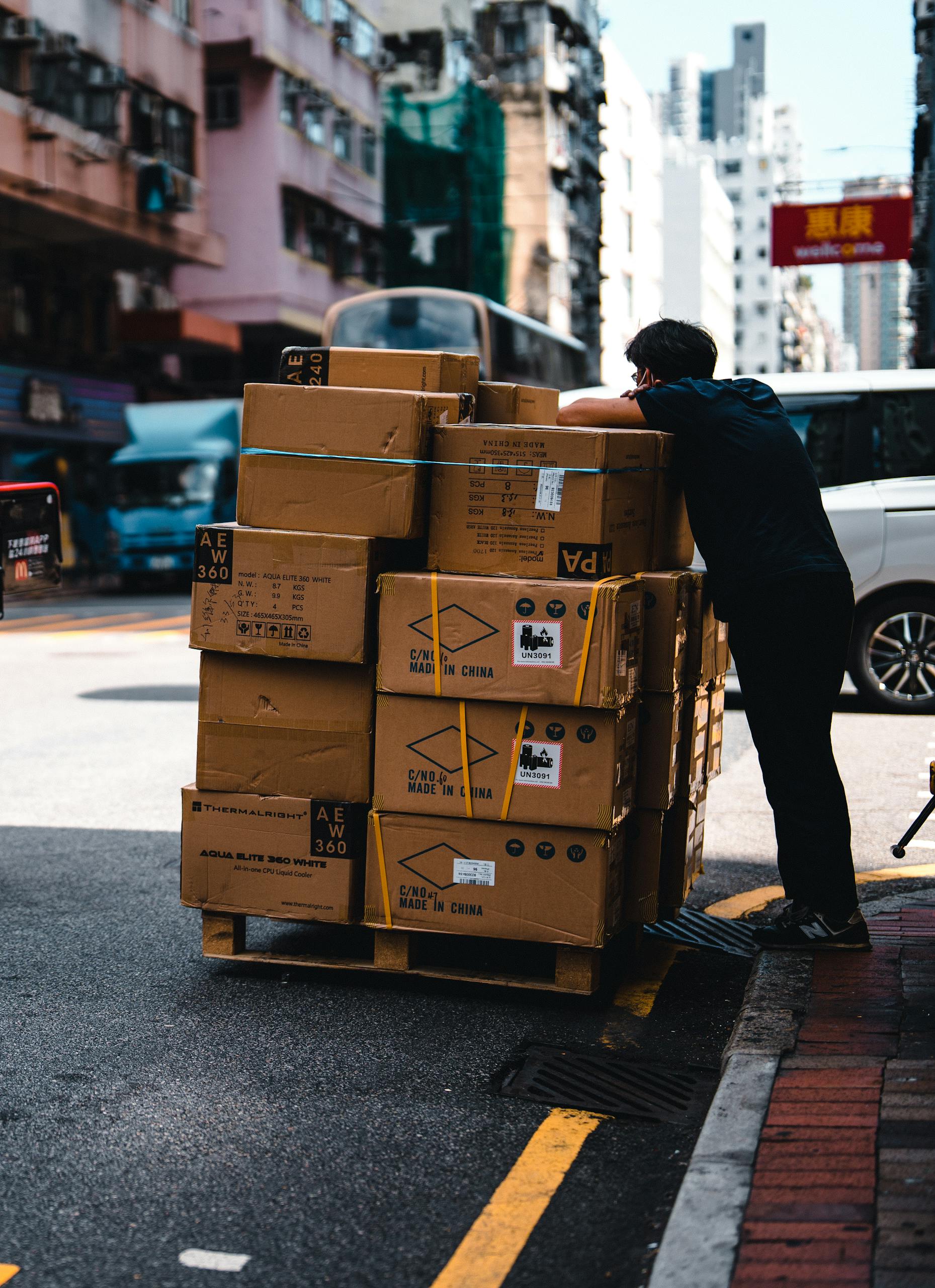 Man handling stacked boxes on a pallet in busy urban street scene.